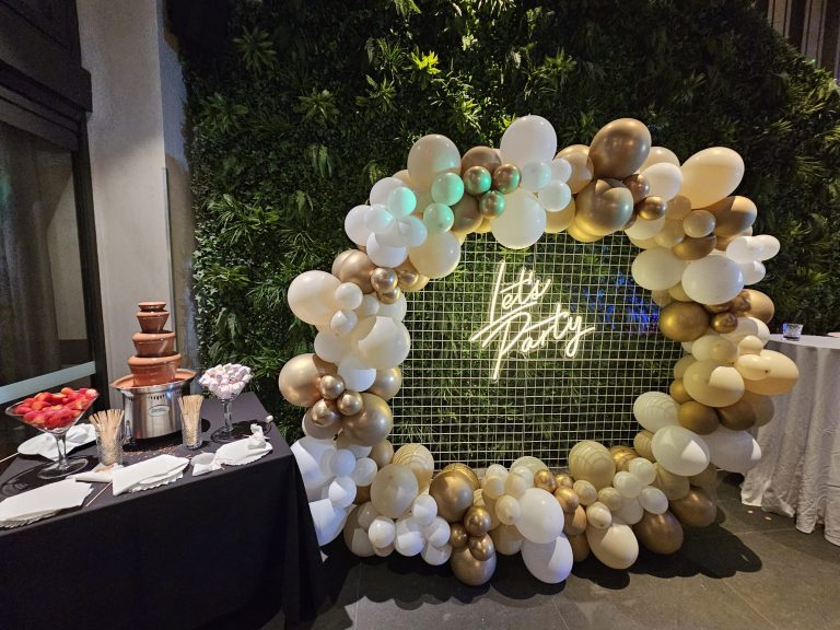 lets party sign attach to a white mesh backdrop with gold and black balloon arrangements. A table with a chocolate fountain